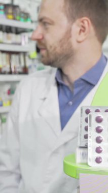 Parlak, modern bir eczanede, odaklanmış bir erkek eczacı dikkatli bir bayan müşteriye, sağlık ve sağlığa olan bağlılığını gösteren, canlı mor haplardan oluşan bir paket sunuyor. Bu ilgi çekici sahne eczacıları vurgular.