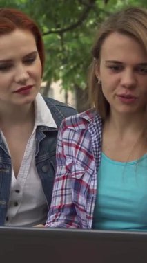 Canlı üç genç öğrenci, iki kadın ve bir erkek, verimli yeşilliklerle çevrili, güneşli bir kampüs bankında dizüstü bilgisayarla neşeyle meşguller. Işıldayan gülümsemeleri ve hareketli ifadeleri dostluk ve işbirliği için davetkar bir ortam yaratıyor.