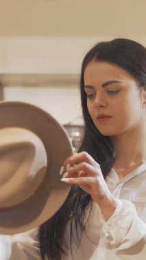 Dalgalı siyah saçlı şık bir kadın butik aynadaki yansımasına hayran kalarak şık bir şapka ayarlıyor. Bu canlı sahne moda keşiflerinin heyecanını yansıtıyor. Özgüvenini ve karşı konulmaz cazibesini vurguluyor.