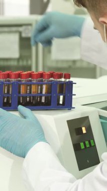 Gıcır gıcır beyaz önlük ve parlak mavi lateks eldivenlerle donanmış çalışkan bir erkek laboratuvar teknisyeni renkli test tüplerini dikkatlice inceleyerek parlak santrifüj için hazırlıyor. Bu büyüleyici sahne, tıbbın titiz doğasını vurguluyor.