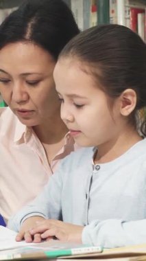 Özenli bir Asyalı anne, sıcak, davetkar bir kütüphanede, hayat dolu kitapların arasında ödevlerini yapmasına yardım ederek tatlı kızına yakın duruyor. Bu dokunaklı an, sevgi ve öğrenme bağını güzel bir şekilde gösteriyor.