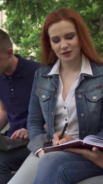 Odaklanmış kahverengi saçlı bir kız öğrenci defterine özenle yazarken, canlı bir kızıl saçlı sınıf arkadaşları hevesle notlar alıyor. Yakınlarda, laptopunda yakışıklı, beyaz bir erkek tip, canlı bir işbirliği ortamı yaratıyor.