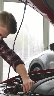 Şık sakallı şık bir genç adam bir arabanın parıldayan kaputunun üzerine eğiliyor, güçlü motoru tatmin olmuş bir gülümsemeyle inceliyor. Modern bayilikler parlak, cilalı arkaplan sahibi olmanın heyecanını artırıyor.