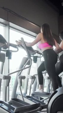 Canlı bir dikiz aynası, geniş pencerelerden süzülen doğal ışığın sıcaklığıyla aydınlatılan eliptik spor ayakkabıları üzerinde enerjik bir şekilde çalışan iki büyük kadını gösteriyor. Neşeli ifadeleri ve ortak motivasyonları güç yayıyor.