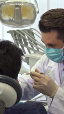 Ağız maskesi ve eldivenleri olan kendini işine adamış genç bir dişçi, müşterilerinin dişlerini parlak, iyi ışıklandırılmış bir diş kliniğinde inceliyor. Odaklanmış Kafkasyalı profesyonel bir diş araştırması yapar, titizlikle eğilir ve eksiksiz bir...