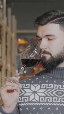 Şık sakallı bir adam zarif bir şekilde bir bardak koyu kırmızı şarabın tadını çıkarıyor, keskin bakışları enfes şişelerle dolu hareketli bir şarap süpermarketinin canlı atmosferini yansıtıyor. Samimi bir süveter giymiş, sofistike ve tutkulu...