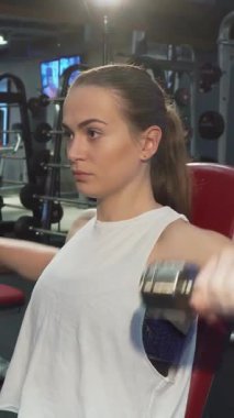 Şık sarı at kuyruklu kararlı genç kadın, yoğun bir şekilde dambılları kaldırıyor etkileyici pazılarını ve sarsılmaz bağlılığını sergiliyor. Yüksek teknolojili spor aletleri ile çevrili. Formda kalma ruhunu somutlaştırıyor. Sınırlarını zorluyor.