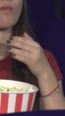 Uzun siyah saçları ve parlak bir gülümsemesi olan neşeli genç bir kadın heyecan verici bir filme dalmışken klasik kırmızı ve beyaz çizgili kovadan patlamış mısırın tadını çıkarıyor. Onun gündelik kırmızı bluzu ve şakacı tırnak cilası bunu canlı bir atmosfere ekliyor.