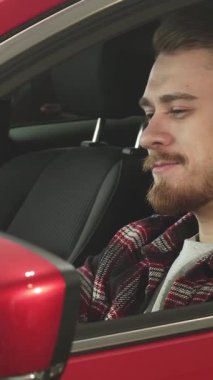 Güzel bakımlı sakallı şık bir genç adam sürücünün koltuğundan neşe saçan göz alıcı kırmızı arabasıyla kendine güveni ve memnuniyeti yayıyor. Rahat duruşu ve büyüleyici gülüşü yeni sahipliğin heyecanını yansıtıyor.