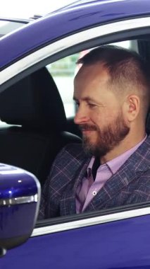 Şık sakallı, şık bir adam, şık bir takım elbise giymiş, parlak mavi bir arabanın sürücü koltuğunda güvenle oturuyor, profesyonellik saçıyor. Çekici gülümsemesi ve şatafatlı iç mekan detaylarını incelemesi coşkusunu vurguluyor.