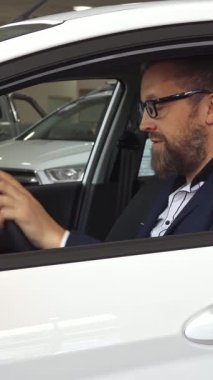 Şık, olgun, bakımlı sakallı beyaz bir adam galeride parlak beyaz bir aracın camından içeri doğru parlak bir araba anahtarı tutarken neşe saçıyor. Çekici gülüşü ve kendine güveni heyecan saçıyor.