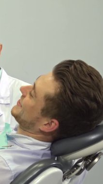 Yakışıklı genç bir adam, şık sarı saçları aynadaki yansımasından dolayı neşeyle parıldıyor dişçi ofisinde göz kamaştırıcı gülümsemesini sergiliyor. Önlük ve maske takmış özenli esmer, beyaz dişçi, diş aletleriyle yakınlarda duruyor.