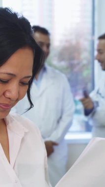 Neşe içinde ışıldayan genç bir kadın, tıbbi test sonuçlarını parlak ve hoş bir ofiste incelerken başparmağıyla onaylıyor. Arka planda, iki erkek doktor düşünceli bir tartışmaya girişirler, destekleyici ve profesyonel bir atmosferi teşvik ederler.