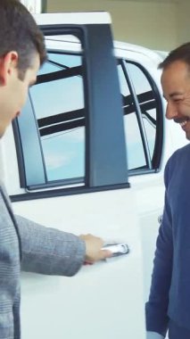 Modern bir araba galerisinde, bakımlı sakallı şık bir satıcı coşkuyla beyaz bir aracın kapısını açar, neşeli bir alıcıyı içeri davet eder. Hava, heyecan ve heyecanla doludur.