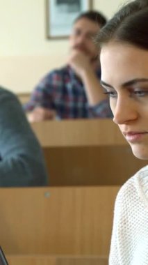 Dalgın bakışlı, meşgul, beyaz bir kız öğrenci parlak, geniş bir konferans salonunda, parlak dizüstü bilgisayarına özenle notlar yazar. Arka planda, iki erkek sınıf arkadaşı derslerine dalmış, biri not defterine düşüncelerini yazıyormuş.