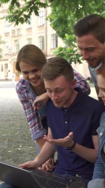 Canlı dört öğrenciden oluşan bir grup, verimli yeşilliklerle çevrili, güneşli bir kampüs gününde dizüstü bilgisayarın etrafında toplanırken kahkahalara boğuluyor. Yakışıklı sarışın adam ve güzel kızıl kız, neşeli sınıf arkadaşları ayakta dikilirken bankta yakın otururlar.