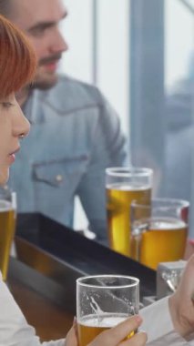 Canlı kızıl saçlı genç bir kadın, hareketli bir barda akıllı telefonuyla takılırken soğuk biranın tadını çıkarıyor. Dostlar toplandıkça, neşeli atmosfer kahkahalarla ve hareketli sohbetlerle uğulduyor.