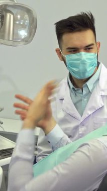 Kendine güvenen koyu renk saçlı genç bir adam, gösterişli, modern bir dişçi koltuğunda rahatça otururken, pozitiflik ve memnuniyet saçarak başparmağını kaldırıyor. Maske ve eldiven takan dişçi profesyonel, yakınlarda duruyor.