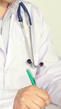 Beyaz önlüklü, odaklanmış beyaz erkek bir doktorun yakın çekimi. Dosyasına canlı yeşil bir kalemle hastanın detaylarını yazıyor. Yumuşak, sıcak aydınlatma sahneyi kaplıyor, steteskopunu vurguluyor ve davetkar bir...