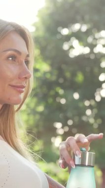 Canlılıkla parlayan ışıl ışıl bir kadın, canlandırıcı açık hava egzersizinden sonra turkuaz şişesinden ferahlatıcı bir yudum su içiyor. Sıcak güneş ışığıyla yıkanmış ve yemyeşil ile çevrili neşeli gülüşü sağlık ve neşe saçıyor.