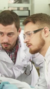 Çıtır beyaz önlükler ve koruyucu eldivenler giymiş iki genç erkek laboratuvar asistanı, bir bilgisayar ekranına dikkatle yaslanmış, test sonuçlarıyla ilgili ayrıntılı bir tartışmaya daldılar. İfadeleri merak ve kararlılık yayar.