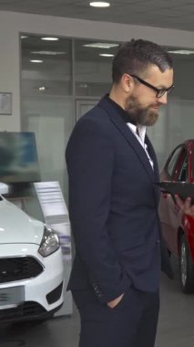 Gösterişli, modern bir araba galerisinde, kendine güvenen siyah takım elbiseli bir satış müdürü, sözleşmenin detaylarını özenli bir müşteriye tutkuyla açıklar. Genç Kafkasyalı danışman parlak bir araca doğru hareket eder, özelliklerini vurgularken,