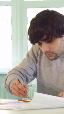 Çekici beyaz bir mimar detaylı bir plan, uzmanlık ve yaratıcılık yayarak kusursuz ölçümler yapmak için kendine güvenen bir pusula kullanır. Sıcak bir gülümseme ile, bu genç erkek tasarımcı güneşli bir masada oturuyor, mimari planlarla çevrili.