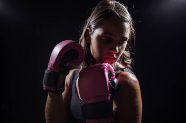 Koyu renk saçlı bir kız, karanlık ve kasvetli bir ortamda kum torbasıyla boks yapıyor. Boks eldiveni giyiyor ve tamamen antrenman veya egzersiz rutinine odaklanmış durumda.. 