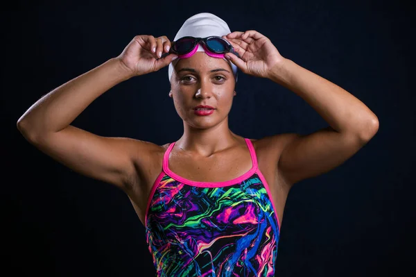 stock image pretty dark haired girl posing in a studio while wearing a training swimwear outfit that includes a swimsuit, swimming goggles, and a swimming cap.