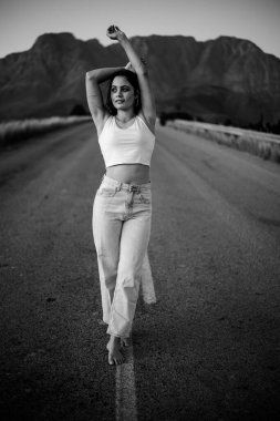 Issız bir yolda siyah saçlı güzel bir genç kadın. O, kot pantolon ve şık bir hasat bluzu da dahil olmak üzere modaya uygun ve şık giyinmiş..