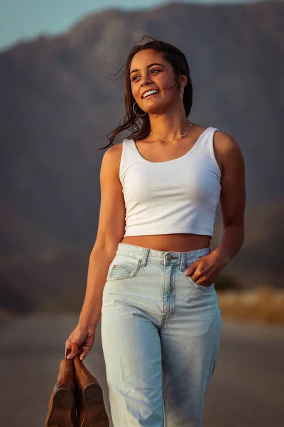 pretty young woman with dark hair on a deserted road. She is dressed in trendy and fashionable attire, including jeans and a stylish crop top.