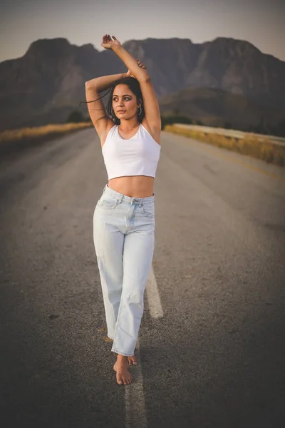 pretty young woman with dark hair on a deserted road. She is dressed in trendy and fashionable attire, including jeans and a stylish crop top.