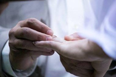 Bu güzel görüntü, bir çiftin gerçek bir düğünde yüzüklerini değiş tokuş ettiği samimi anı yakalıyor. Fotoğrafta çiftin ellerinin birbirine dolanmış parmaklarını ve parmaklarındaki alyansları gösteren yakın çekimi yer alıyor. Görüntü