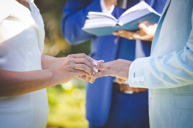 Bu güzel görüntü, bir çiftin gerçek bir düğünde yüzüklerini değiş tokuş ettiği samimi anı yakalıyor. Fotoğrafta çiftin ellerinin birbirine dolanmış parmaklarını ve parmaklarındaki alyansları gösteren yakın çekimi yer alıyor. Görüntü