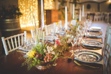 Bu büyüleyici görüntü, gerçek bir düğünün zarif dekorunu ve büyüleyici çiçek düzenlemelerini gözler önüne seriyor. Fotoğrafta zarif çiçekler, mumlar ve diğer dekoratif el ile süslenmiş büyüleyici bir düğün yerinde güzel dekore edilmiş bir masa yer alıyor.