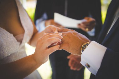 Bu güzel görüntü, bir çiftin gerçek bir düğünde yüzüklerini değiş tokuş ettiği samimi anı yakalıyor. Fotoğrafta çiftin ellerinin birbirine dolanmış parmaklarını ve parmaklarındaki alyansları gösteren yakın çekimi yer alıyor. Görüntü