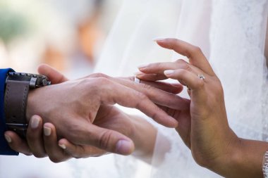 Bu güzel görüntü, bir çiftin gerçek bir düğünde yüzüklerini değiş tokuş ettiği samimi anı yakalıyor. Fotoğrafta çiftin ellerinin birbirine dolanmış parmaklarını ve parmaklarındaki alyansları gösteren yakın çekimi yer alıyor. Görüntü