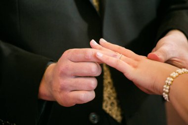 Bu güzel görüntü, bir çiftin gerçek bir düğünde yüzüklerini değiş tokuş ettiği samimi anı yakalıyor. Fotoğrafta çiftin ellerinin birbirine dolanmış parmaklarını ve parmaklarındaki alyansları gösteren yakın çekimi yer alıyor. Görüntü
