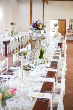 Bu büyüleyici görüntü, gerçek bir düğünün zarif dekorunu ve büyüleyici çiçek düzenlemelerini gözler önüne seriyor. Fotoğrafta zarif çiçekler, mumlar ve diğer dekoratif el ile süslenmiş büyüleyici bir düğün yerinde güzel dekore edilmiş bir masa yer alıyor.