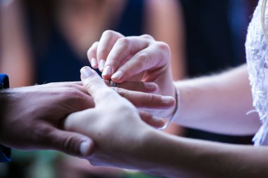 Bu güzel görüntü, bir çiftin gerçek bir düğünde yüzüklerini değiş tokuş ettiği samimi anı yakalıyor. Fotoğrafta çiftin ellerinin birbirine dolanmış parmaklarını ve parmaklarındaki alyansları gösteren yakın çekimi yer alıyor. Görüntü
