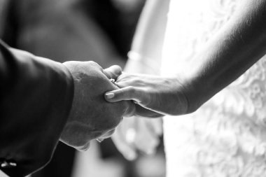 Bu güzel görüntü, bir çiftin gerçek bir düğünde yüzüklerini değiş tokuş ettiği samimi anı yakalıyor. Fotoğrafta çiftin ellerinin birbirine dolanmış parmaklarını ve parmaklarındaki alyansları gösteren yakın çekimi yer alıyor. Görüntü