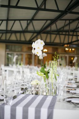 Bu büyüleyici görüntü, gerçek bir düğünün zarif dekorunu ve büyüleyici çiçek düzenlemelerini gözler önüne seriyor. Fotoğrafta zarif çiçekler, mumlar ve diğer dekoratif el ile süslenmiş büyüleyici bir düğün yerinde güzel dekore edilmiş bir masa yer alıyor.