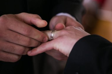 Bu güzel görüntü, bir çiftin gerçek bir düğünde yüzüklerini değiş tokuş ettiği samimi anı yakalıyor. Fotoğrafta çiftin ellerinin birbirine dolanmış parmaklarını ve parmaklarındaki alyansları gösteren yakın çekimi yer alıyor. Görüntü