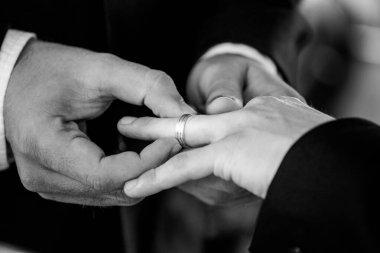 Bu güzel görüntü, bir çiftin gerçek bir düğünde yüzüklerini değiş tokuş ettiği samimi anı yakalıyor. Fotoğrafta çiftin ellerinin birbirine dolanmış parmaklarını ve parmaklarındaki alyansları gösteren yakın çekimi yer alıyor. Görüntü