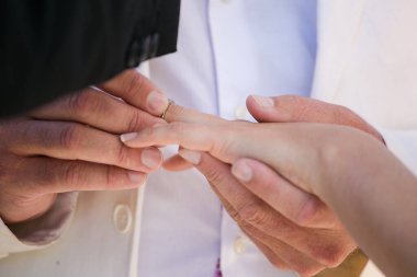 Bu güzel görüntü, bir çiftin gerçek bir düğünde yüzüklerini değiş tokuş ettiği samimi anı yakalıyor. Fotoğrafta çiftin ellerinin birbirine dolanmış parmaklarını ve parmaklarındaki alyansları gösteren yakın çekimi yer alıyor. Görüntü