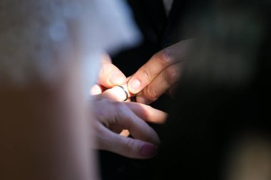 Bu güzel görüntü, bir çiftin gerçek bir düğünde yüzüklerini değiş tokuş ettiği samimi anı yakalıyor. Fotoğrafta çiftin ellerinin birbirine dolanmış parmaklarını ve parmaklarındaki alyansları gösteren yakın çekimi yer alıyor. Görüntü