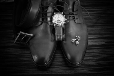 Bu büyüleyici görüntü, damadın büyük günü için giyinirken takım elbisesi, ayakkabıları ve kol düğmeleri de dahil olmak üzere düğün kıyafetini gösteriyor. Fotoğraf, damadın kıyafetinin karmaşık detaylarını vurguluyor. Güzel bir kompozisyon oluşturuyor. 