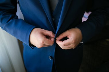 Bu büyüleyici görüntü, damadın büyük günü için giyinirken takım elbisesi, ayakkabıları ve kol düğmeleri de dahil olmak üzere düğün kıyafetini gösteriyor. Fotoğraf, damadın kıyafetinin karmaşık detaylarını vurguluyor. Güzel bir kompozisyon oluşturuyor. 