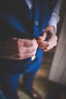 Bu büyüleyici görüntü, damadın büyük günü için giyinirken takım elbisesi, ayakkabıları ve kol düğmeleri de dahil olmak üzere düğün kıyafetini gösteriyor. Fotoğraf, damadın kıyafetinin karmaşık detaylarını vurguluyor. Güzel bir kompozisyon oluşturuyor. 