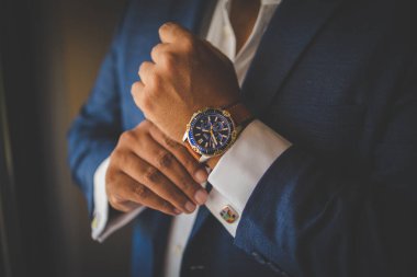 Bu büyüleyici görüntü, damadın büyük günü için giyinirken takım elbisesi, ayakkabıları ve kol düğmeleri de dahil olmak üzere düğün kıyafetini gösteriyor. Fotoğraf, damadın kıyafetinin karmaşık detaylarını vurguluyor. Güzel bir kompozisyon oluşturuyor. 